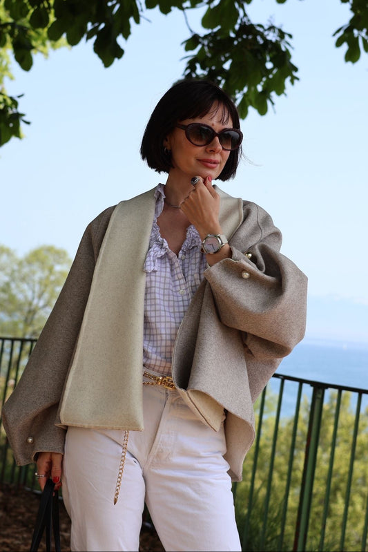 Woman in beige coat and white pants standing outdoors with trees and sky in the background